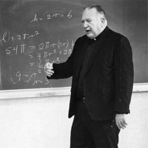 Father Clarence J. Wallen teaching a class in front of a chalkboard in 1985